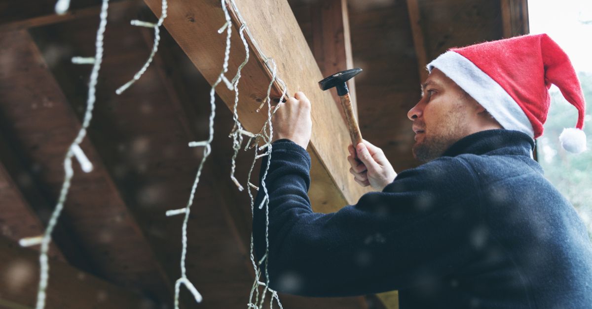 Man in a Santa hat nailing holiday lights to his house