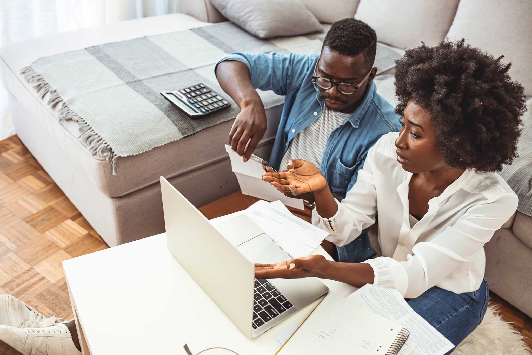 Image of a black man and black woman, working on a laptop computer. A calculator sits in the back ground. The couple looks upset, possibly victims of banking discrimination